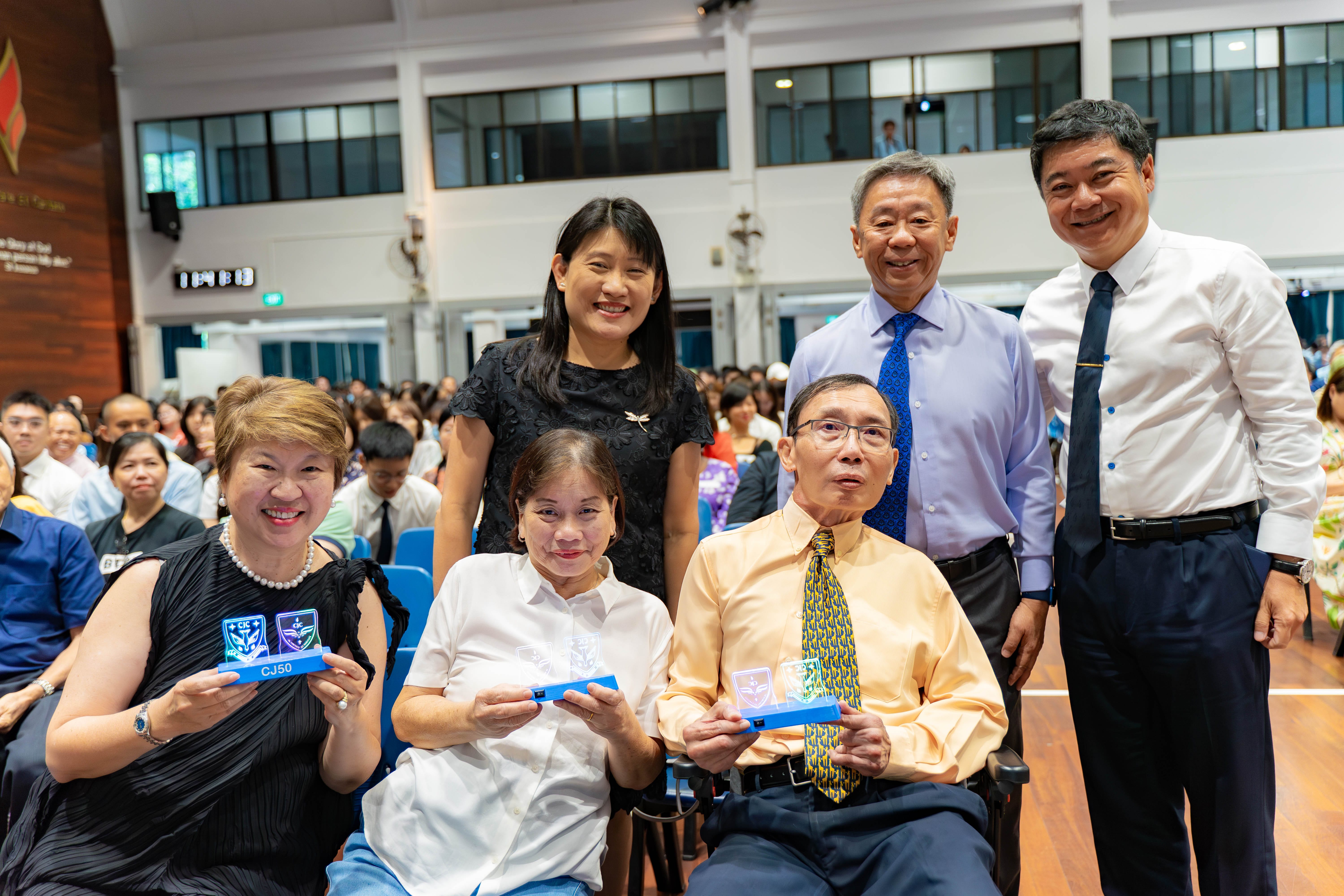 Six people pose together in a hall, with two seated holding blue award boxes labeled CJ50, and four standing behind them.